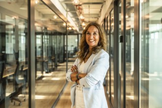 Mature businesswoman standing with folded arms in a bright modern office corridor, exuding