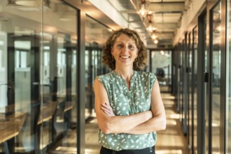 Professional woman with curly hair and arms crossed, confidently standing in a well lit modern