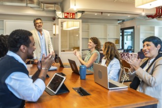 Diverse business colleagues sitting around a large wooden table, smiling and clapping for a speaker