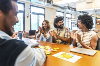 Diverse business colleagues sitting at a table in a modern office, happily clapping and smiling