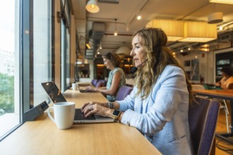 Woman with a headset and microphone actively working on a laptop at a long table by a window,
