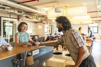 Young man passing a cup of coffee to a smiling woman working on her laptop at a counter in a