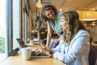 Two women wearing headsets are working together at a desk by a window, with one colleague providing