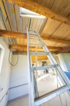 View of an attic with a ladder leading to an open window. Wooden ceiling elements and sunlight