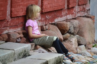 Little blonde girl sitting next to a red half-timbered house and looking to the side in Den gamle