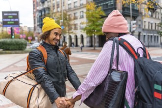 Multiethnic gay couple holding hands, beanies and backpacks, smiling as they walk through a vibrant