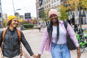 Young multiracial couple smiling and holding hands while strolling down a city street lined with