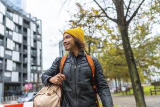 Young man with a yellow beanie and black leather jacket carrying a duffel bag and backpack, smiling