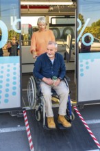 Senior woman is confidently assisting a mature man in a wheelchair to exit a public bus using the