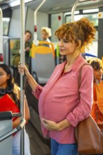 Pregnant woman stands attentively inside a public bus, holding a handrail and gently touching her