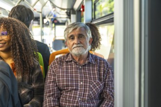 Senior man with a gray beard and plaid shirt sitting on a public bus, commuting with other diverse