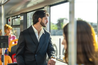 Young businessman in a dark suit stands on a city bus, gazing out the window as warm sunlight