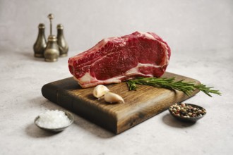 Raw ribeye roll steak on a wooden cutting board surrounded by garlic cloves, fresh rosemary, salt,