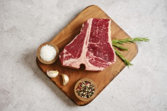 A large, marbled raw t-bone steak on a wooden cutting board surrounded by garlic cloves, salt,