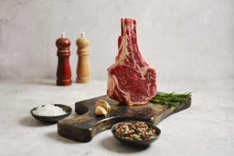 A raw cowboy steak on a wooden cutting board surrounded by fresh herbs. In the background, salt and