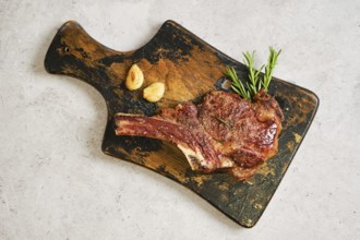 Grilled cowboy steak on a worn wooden cutting board, garnished with fresh rosemary and garlic. The