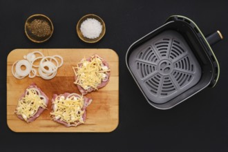 Raw pork chops are topped with grated cheese and sliced onions on a wooden cutting board. Nearby,