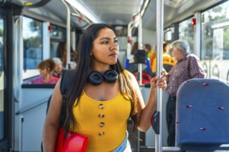 Young woman passenger holding a handrail inside a public bus, wearing headphones and a backpack,