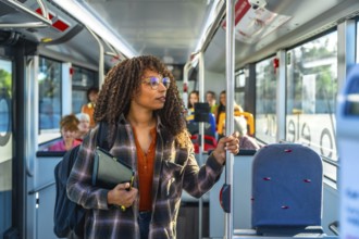 Young woman with curly hair and glasses stands inside a city bus holding a pole, wearing a backpack