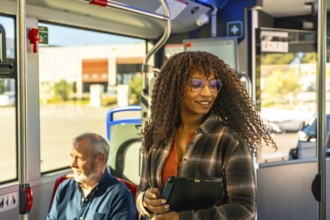 Woman with glasses smiles while standing on a city bus, holding a digital tablet among commuters