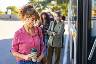 Smiling woman holding a reusable coffee cup waits in line at a busy city bus stop with diverse