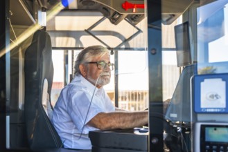 Senior bus driver with eyeglasses and white shirt focused on steering from the cabin, demonstrating