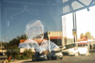 Senior bus driver smiling while looking outside through a window, reflecting street traffic,