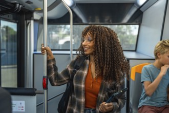 Woman with glasses and curly hair holding a digital tablet while commuting on a public bus, smiling