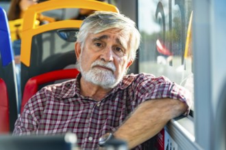 Senior man with white beard and plaid shirt sits by a bus window, gazing pensively at the camera