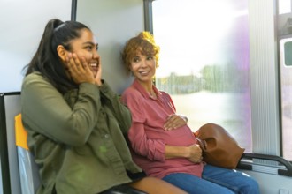 Young woman is smiling and showing surprise while looking at her pregnant friend, who is gently