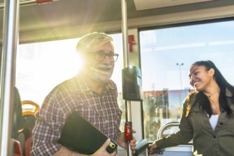 Two smiling passengers, an older man and a young woman, are traveling on a bus during daylight,