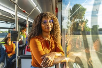 Smiling black woman with curly hair and glasses stands on a city bus during her commute, confident
