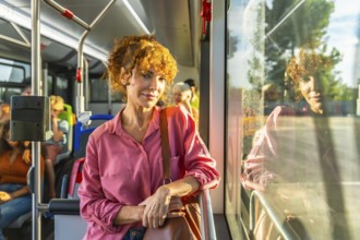 Young redheaded woman smiling and looking at camera while standing on a city bus, holding a bag