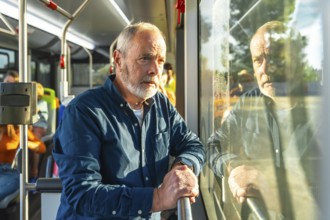 Senior bearded man standing on a city bus, holding a handrail and gazing out the window, bathed in