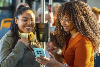 Two smiling multiethnic women bonding and sharing content on a smartphone while commuting together