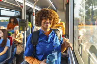 Young nurse in blue scrubs and backpack smiles at camera while standing on a city bus, holding a