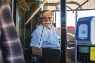 Mature man working as a bus driver, wearing glasses and a white uniform shirt, sitting inside the