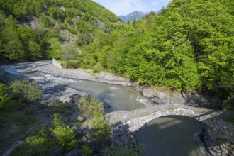 A clear river flows under an old stone bridge through a green, wooded valley with mountains in the