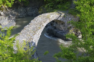 An old stone bridge spans a river amid green trees and rocks, Purtio stone arch bridge,