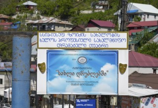 A large advertising sign with a blue poster and text surrounded by colorful buildings and green