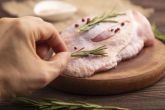 Raw Turkey Thigh with spices and rosemary on cutting board with hand on brown wooden background and