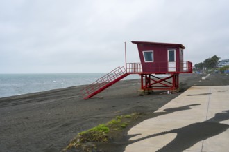 Red painted rescue tower stands abandoned on cloudy beach, beach with magnetic sand, magnetite,