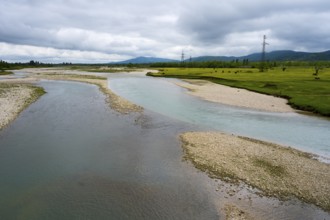 A river snakes through strips of pebbles flanked by green fields and cloudy skies, interrupted by