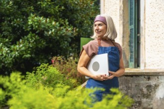 Senior woman in apron and headband holding a laptop outdoors in her garden, smiling and looking