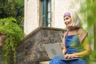 Senior woman enjoying remote work from a peaceful garden setting, smiling while typing on a laptop,