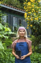Senior woman wearing an apron and a headband, standing in a lush green garden while holding a