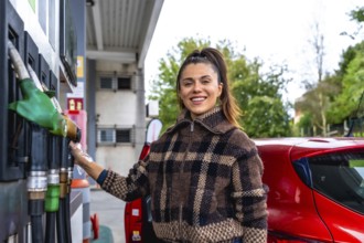 Smiling young woman in a plaid jacket and protective glove refuels her red car at an urban gas