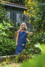 Senior woman in an apron and headband standing in a lush home vegetable garden, smiling while