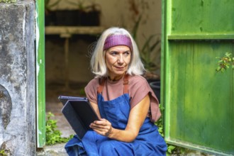 Senior woman artist in an apron and headband working on a digital tablet outside a rustic green