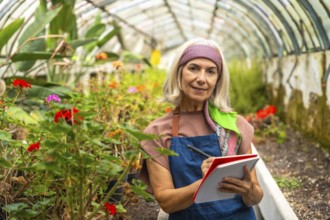 Senior woman wearing an apron and headband smiling, standing inside a botanical greenhouse while